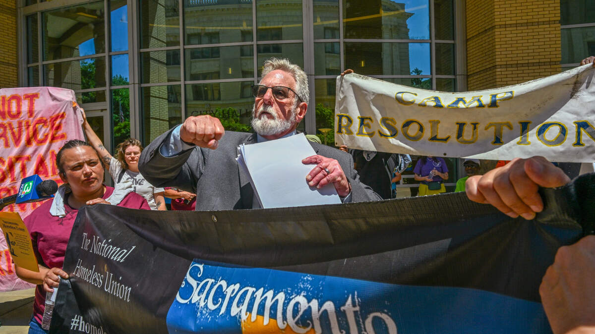 El abogado Anthony Prince levanta el puño en una rueda de prensa celebrada el miércoles en el Ayuntamiento de Sacramento mientras muestra los documentos de una demanda que tiene previsto presentar ante la Corte Superior de Sacramento en la que alega que la ciudad está incumpliendo su contrato de arrendamiento con Camp Resolution, un campamento autónomo de personas sin hogar situado en el norte de Sacramento. "Camp Resolution es un faro de lucha, un faro de esperanza, un faro de organización", dijo.