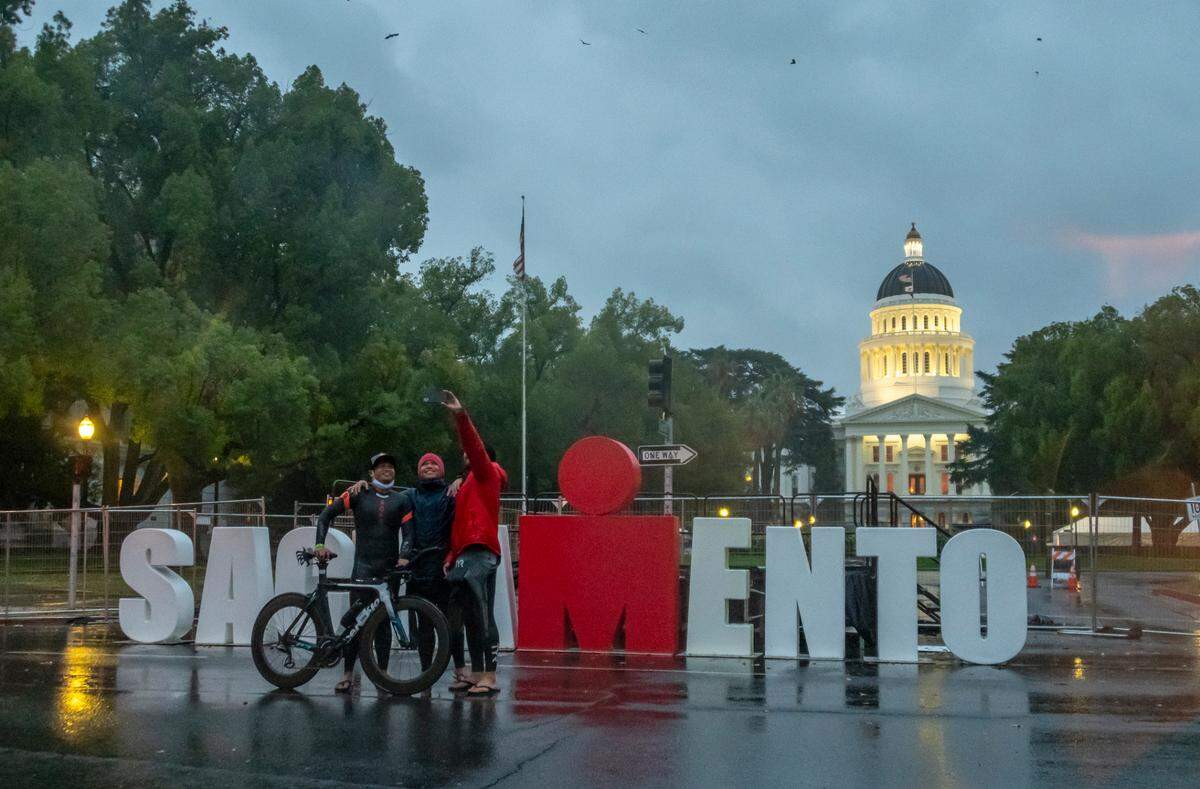 Orange County residents Raymond Soquena, Gary Binoya and Mike Sacramone, who had all planned to participate in the Ironman triathlon, take a selfie in front of the podium at the planned finish after the race was canceled because of weather on Sunday, Oct. 24, 2021.