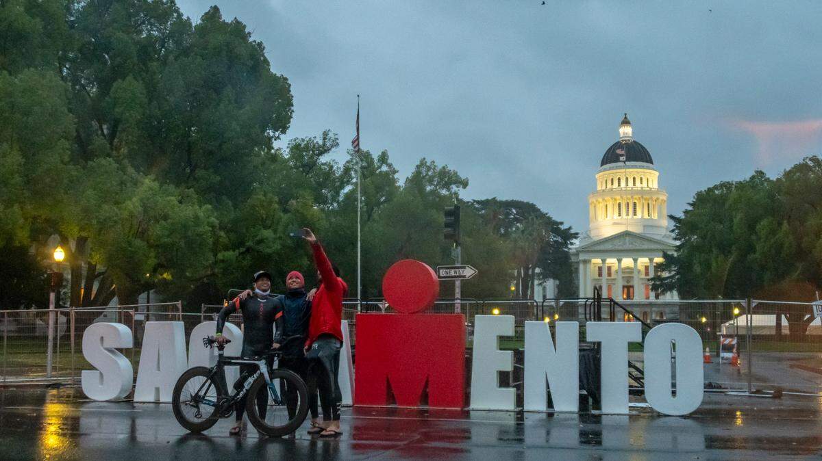 Orange County residents Raymond Soquena, Gary Binoya and Mike Sacramone, who had all planned to participate in the Ironman triathlon, take a selfie in front of the podium at the planned finish after the race was canceled because of weather on Sunday, Oct. 24, 2021.