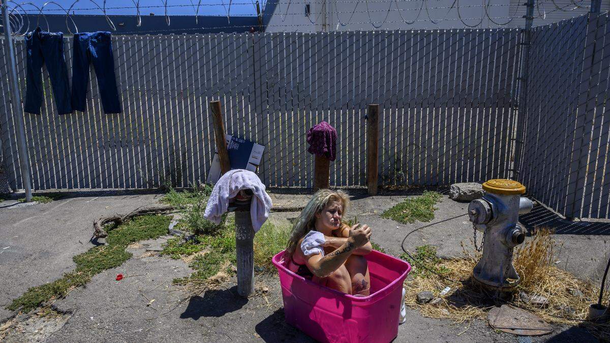 Laurane Ivey, 37, sinks deep into a pink plastic tub filled with water from a fire hydrant as she begins to scrub the dirt from her worn feet. Above her is a barbed wire fence surrounding the shelter she used to live in before it closed. With no place to go, she lives in her car, joining other people experiencing homelessness and living in parked cars across the street, including her mom Gwen Mayes, 59, who sleeps with two tiny dogs for protection. She says she wished there was a designated parking spot for the homeless with showers and portable toilets. She says she has to go to the bathroom in a bag inside her car.