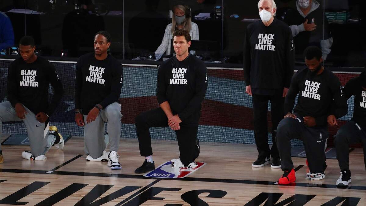 San Antonio Spurs head coach Gregg Popovich, fourth from left, stands while wearing a mask while Sacramento Kings head coach Luke Walton, center, kneels with players their game Friday night.