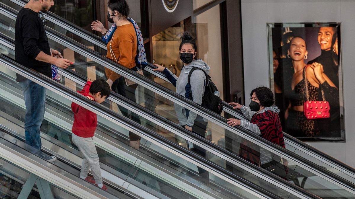 Shoppers, some with masks and some without, ride the escalator at Sacramento’s Arden Fair mall near an advertisement of smiling models outside the Michael Kors store on Tuesday, Feb. 15, 2022.