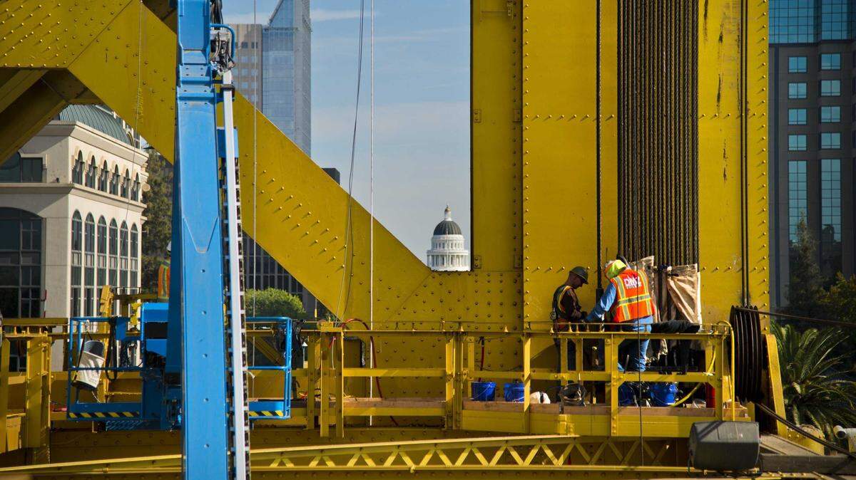 Two construction workers stand on a platform beside the lift cables of the Tower Bridge in Sacramento in 2018. Caltrans is conducting a $3 million project that includes electrical work and component replacements, the first major work on the bridge since crews replaced its aging lift cables eight years ago.