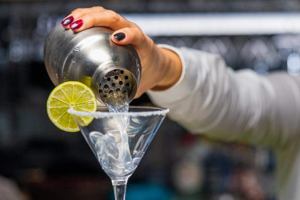 A female bartender pours a margarita drink into a glass from a stainless steel cocktail shaker.