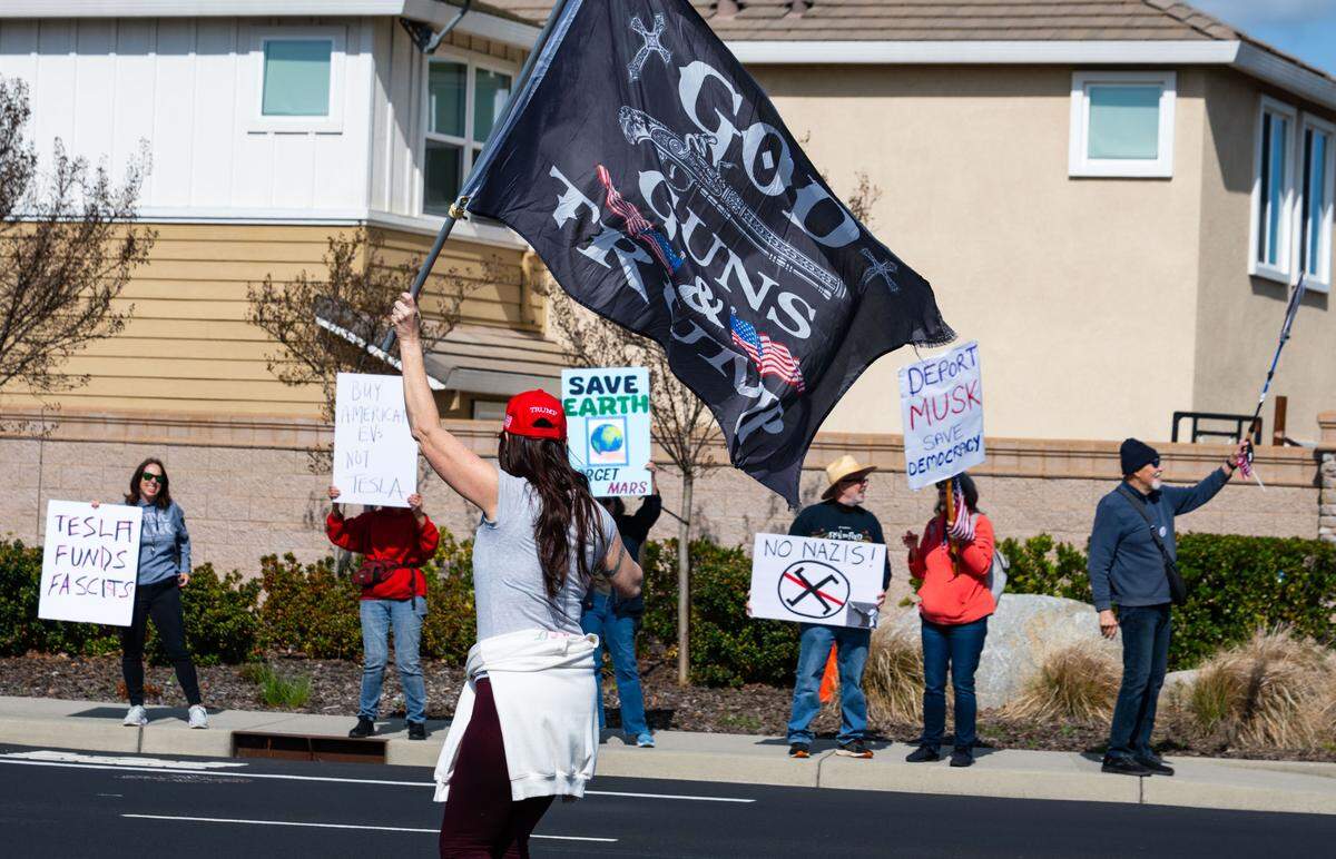A woman with a flag that says “God, guns and Trump” marches down the center of Granite Drive in Rocklin across from the Tesla dealership as anti-Tesla protesters hold signs on Saturday, March 29, 2025.