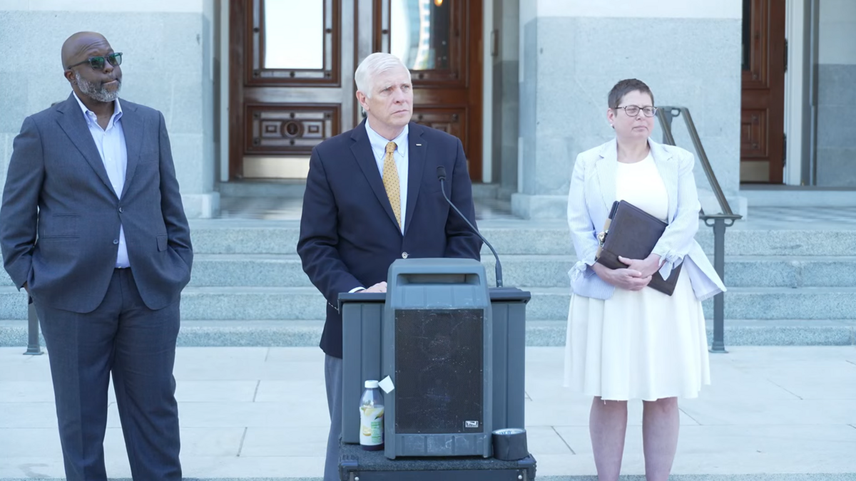 State Sen. Tom Umberg stands at the podium between Darius Kemp (left) and Dora Rose (right) at “Hands Off our Elections” press conference on Thursday, June 11, 2026, at the Capitol in Sacramento.
