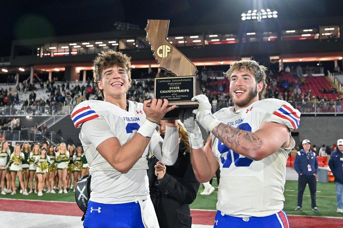 Folsom Bulldogs quarterback Ryder Lyons (3) and defensive tackle Lucas Hardeman (56) hold the state championship plaque after winning the 2023 CIF Division 1-A state championship against the St. Bonaventure Seraphs. The program's success gives it an advantage in finding the latest drills to teach technique and avoid injuries. 