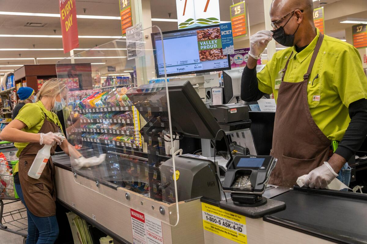 Cashier Paul Watkins, right, adjusts his mask behind a safety shield at Save Mart supermarket in East Sacramento while waiting for Kylie Lenzi, service specialist, left, who cleans the counter between customers, Wednesday, April 8, 2020.