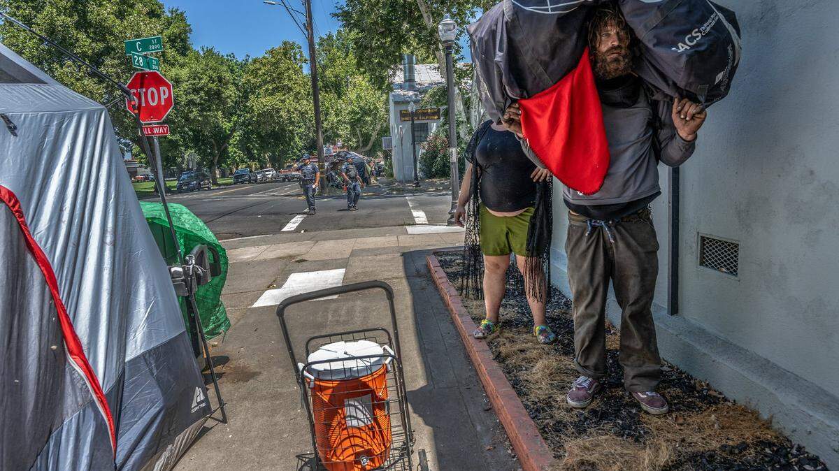 Homeless camper Chadwick Justin Foy, 41, holds all his belongings on his shoulders as friend Valencia Hardwich, 35, steps behind him in the shade while Sacramento police patrol the intersection of 28th and C streets on Tuesday, July 18, 2023. On Tuesday, the Sacramento City Council voted 5-4 to authorize legalized homeless encampments in the city. Leaders hope this will create more capacity to get people off the streets and into safe environments.
