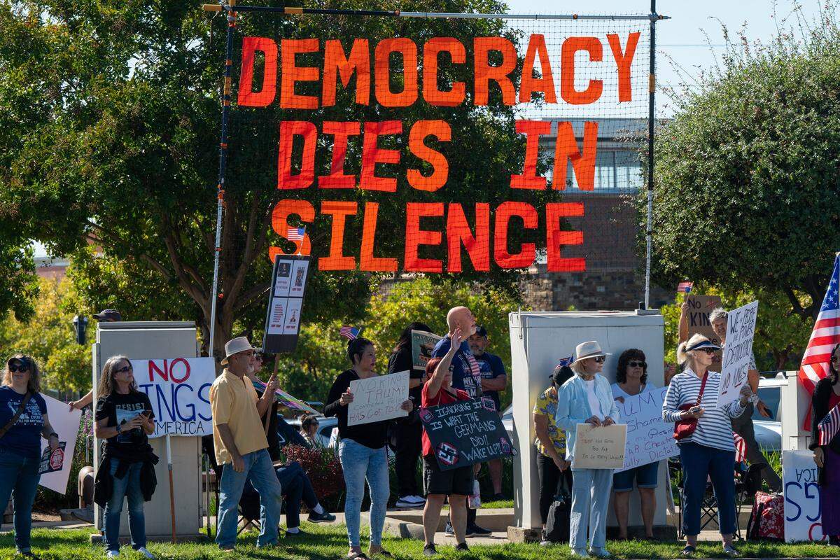 Protesters participate in a “No Kings” rally below a sign that says "Democracy dies in silence" in Roseville on Saturday, Oct. 18, 2025.