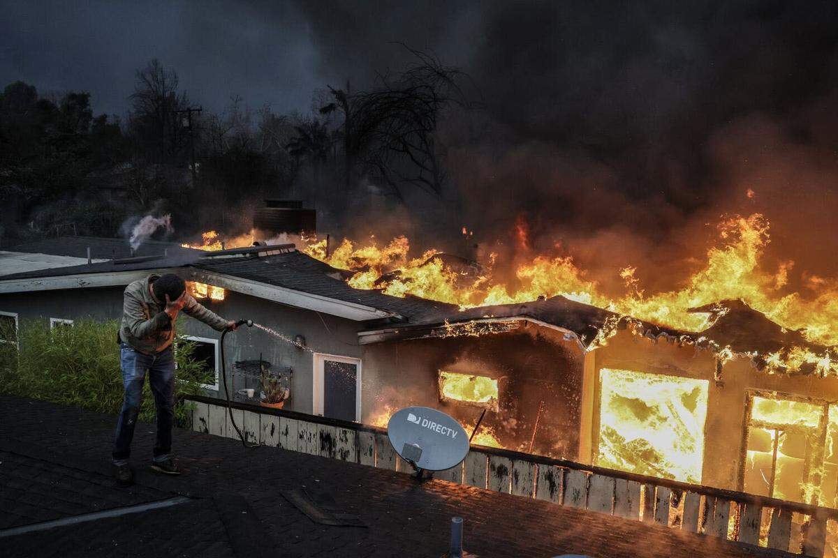 Steve Salinas shields himself from intense heat as he hoses down a neighbors’ rooftop on Sinaloa Avenue as the Eaton Fire burns in Altadena on Wednesday, Jan. 8, 2025.