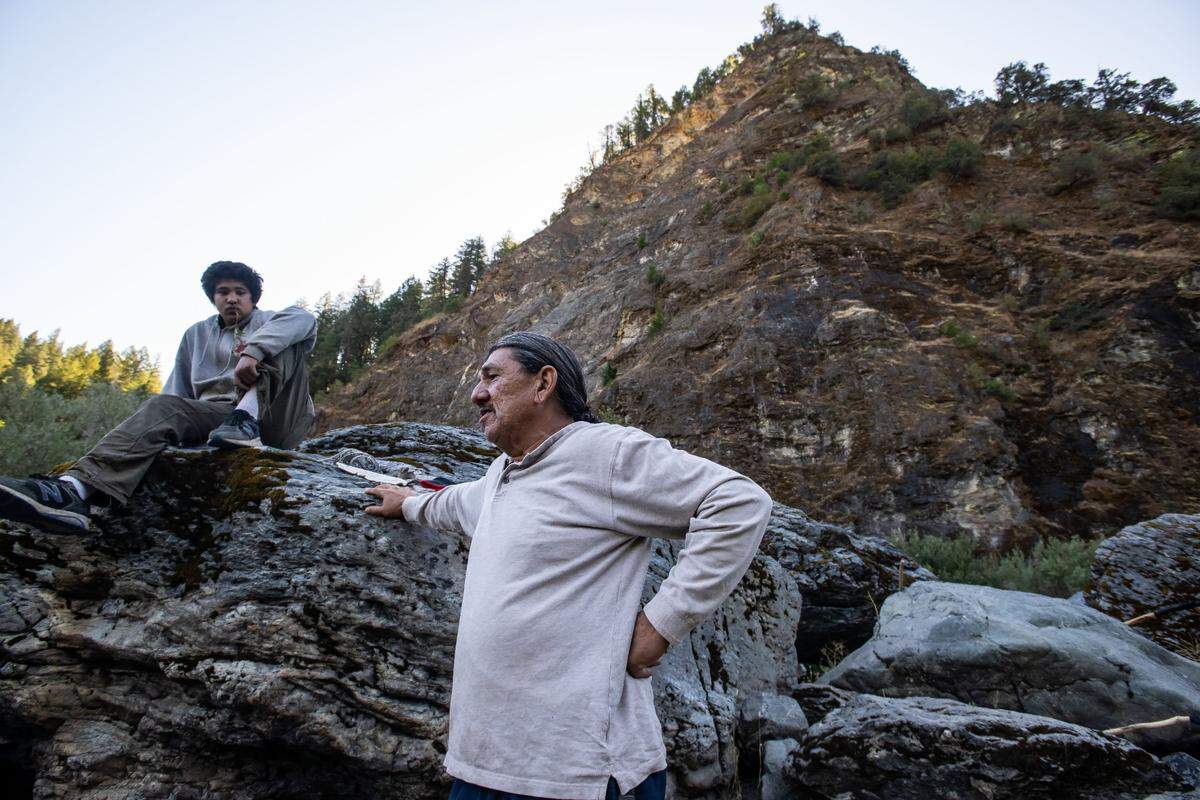 Ron Reed, right, a Karuk Tribe fisherman, talks with fellow tribal members as 13-year-old Ishikiihara E-kor, son of medicine man Chook-Chook Hillman, listens from a rock below the peak of Á’uuyich near Ishi Pishi Falls in late September in Siskiyou County. Á’uuyich is the Karuk’s center of the universe, Reed said.