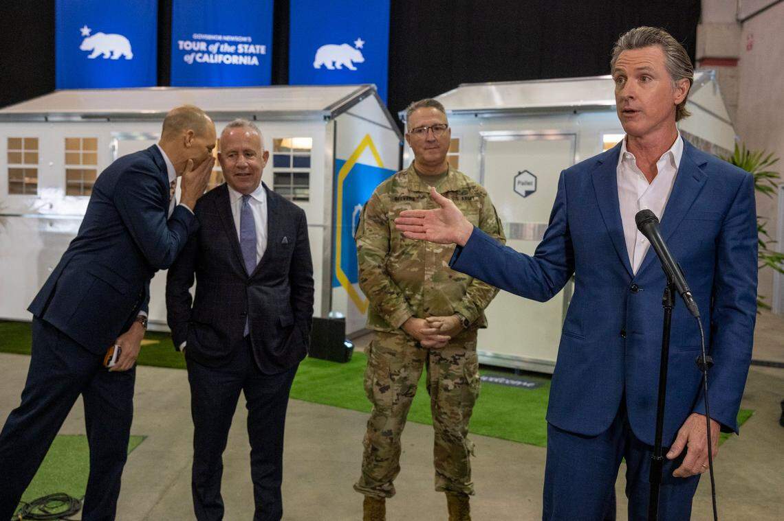 After touring tiny homes at Cal Expo, Gov. Gavin Newsom answers questions from the media March 16, 2023, after announcing that the state will have 1,200 of the homes built and delivered throughout California to help house the homeless population. Assemblyman Kevin McCarty, D-Sacramento, whispers to Sacramento Mayor Darrell Steinberg at left, as California National Guard Maj. Gen. Matt Beevers listens.