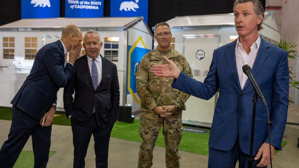 After touring tiny homes at Cal Expo, Gov. Gavin Newsom answers questions from the media on Thursday, March 16, 2023, after announcing that the state will have 1,200 of the homes built and delivered throughout California to help house the homeless population. Assemblyman Kevin McCarty, D-Sacramento, whispers to Sacramento Mayor Darrell Steinberg at left, as California National Guard Maj. Gen. Matt Beevers listens.