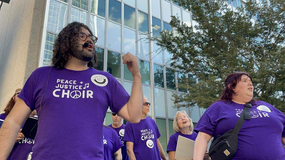 Peace and Justice Choir Director Jason Block directs Kit Rosander, right, Mark Tavianini, JoAnn Fuller, both in background, and other singers in their performance at a Thursday, Oct. 9, 2025, protest.
