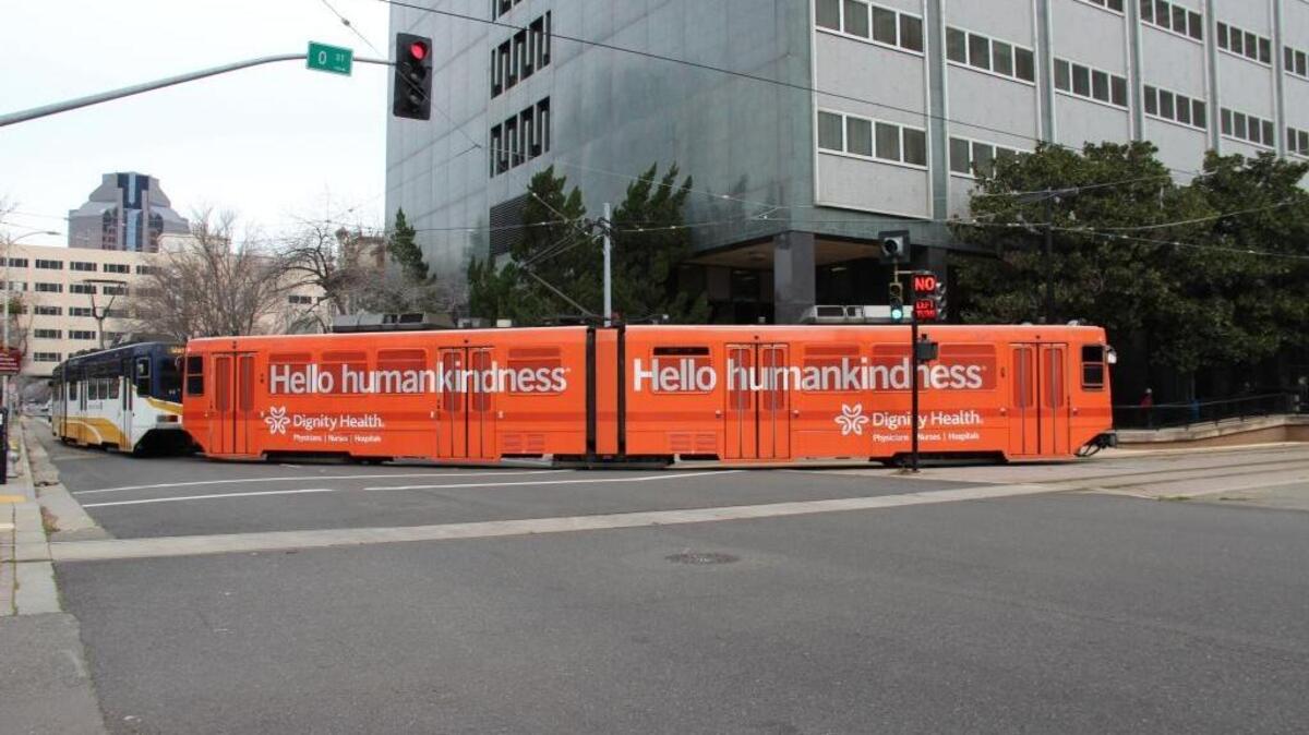 A light-rail train rolls through Sacramento, decked out with a colorful advertisement for Dignity Health. Various advertisements have popped up on light-rail cars this year as part of a recently formed partnership between the Sacramento Regional Transit District and Lamar Advertising Co., a national advertising firm based in Baton Rouge, La. The displays are made of vinyl material “and are applied like a giant bumper sticker.”