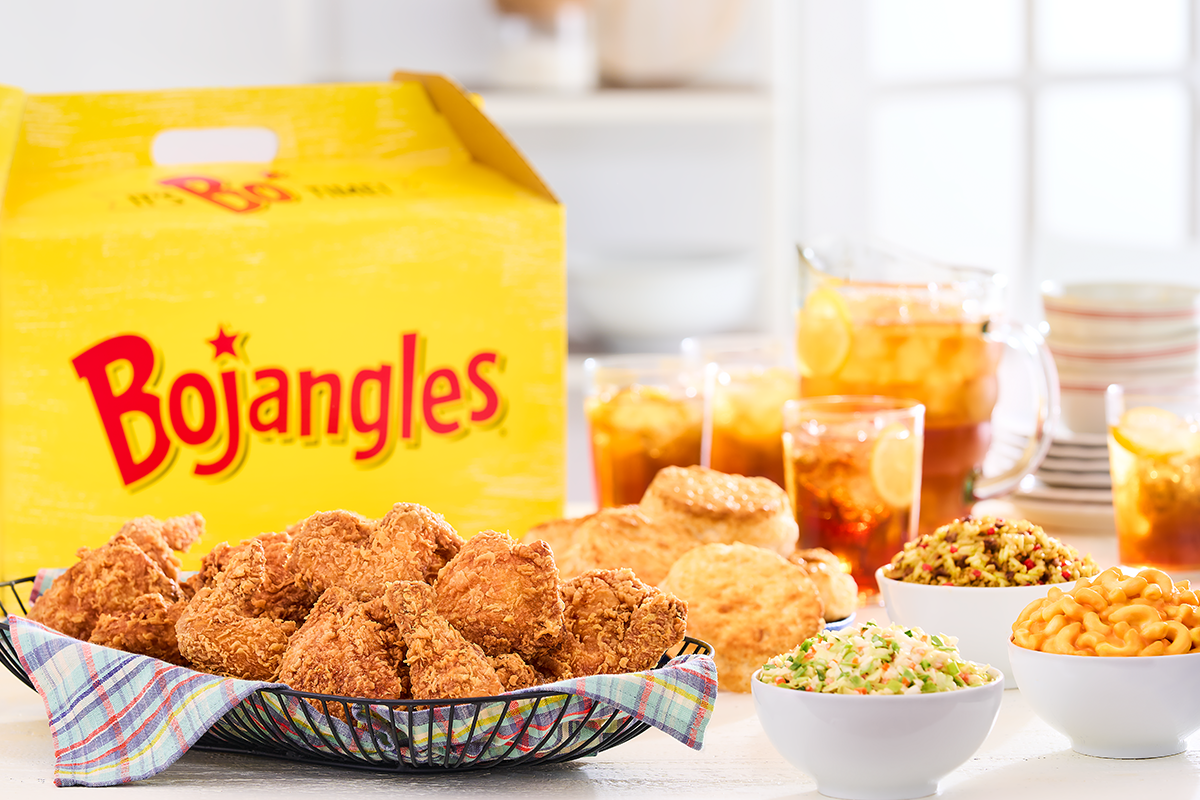 A full meal from Bojangles’ is shown with a large yellow box behind a spread of food. A basket of fried chicken is in the foreground, with biscuits, iced tea, and side dishes of macaroni and cheese, rice, and coleslaw arranged around it.