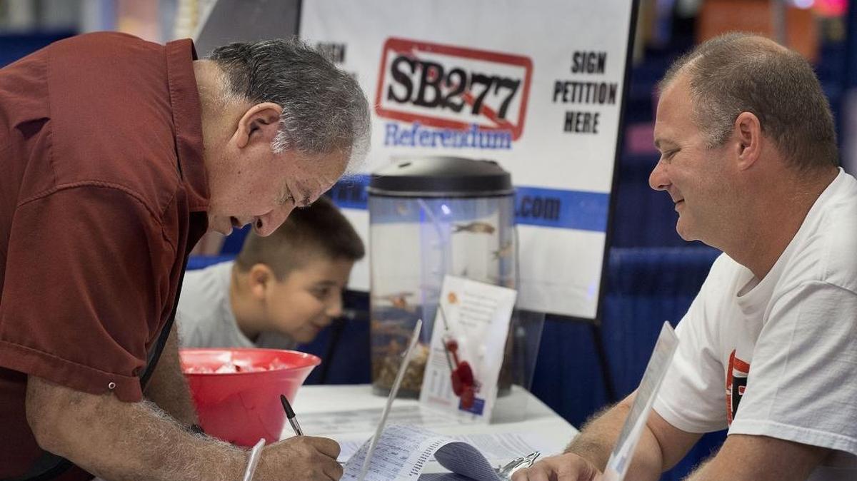 
Adam Cobarrubio of Stockton, left, with his grandson Andrew Cobarrubio, 9, signs a petition at a booth at the Lodi Grape Festival in Lodi on Friday, Sept. 18, 2015. A group had a booth at the festival to garner signatures to qualify a referendum on SB 277, the law requiring schoolchildren to get vaccinated. Andrew is peering into a fish tank filled with 50 goldfish – equal to the number of vaccine injections mandated for children up to age 18.
