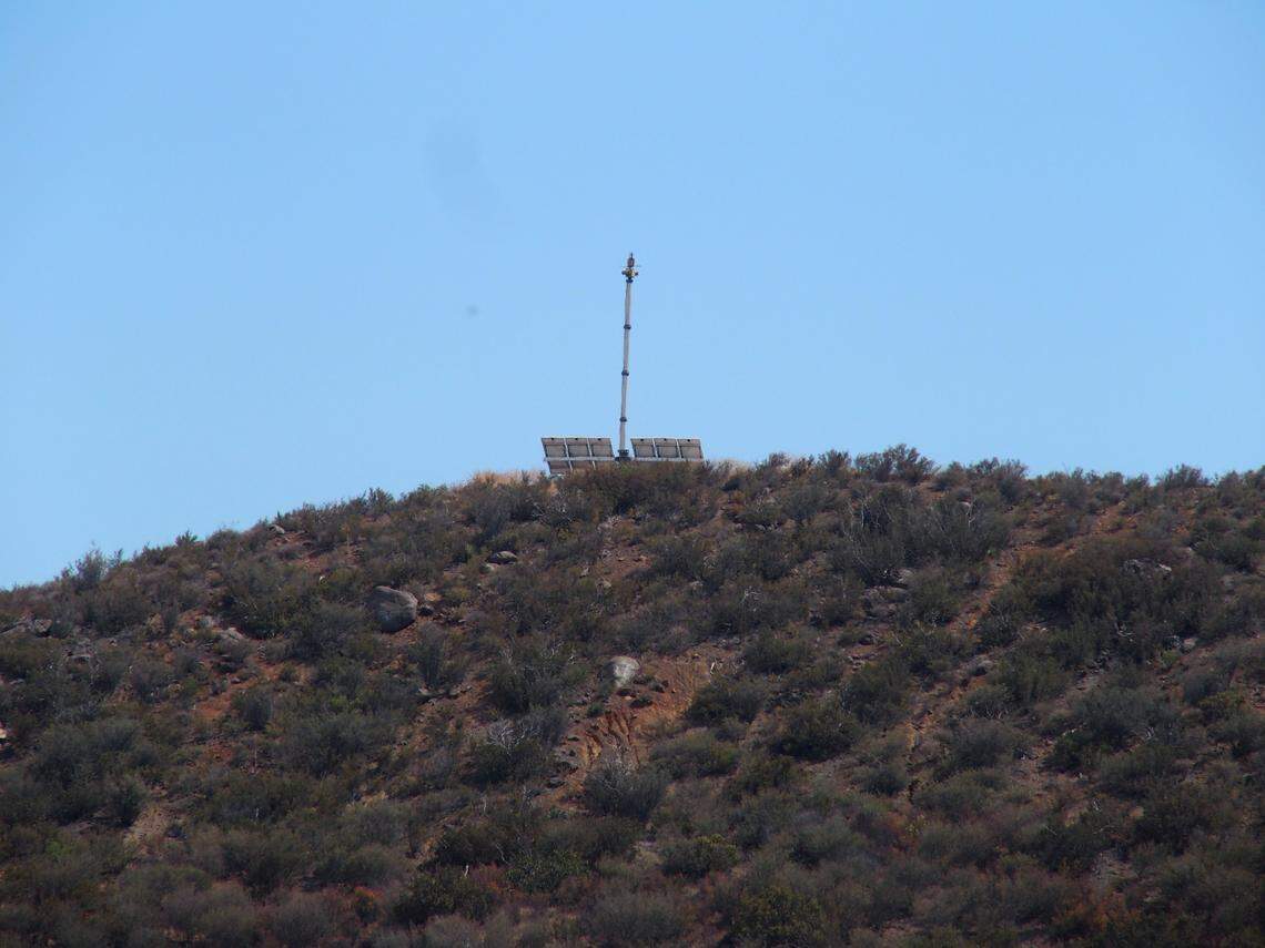 An Anduril Sentry surveillance device perches on a low hill covered in scrub and bushes.