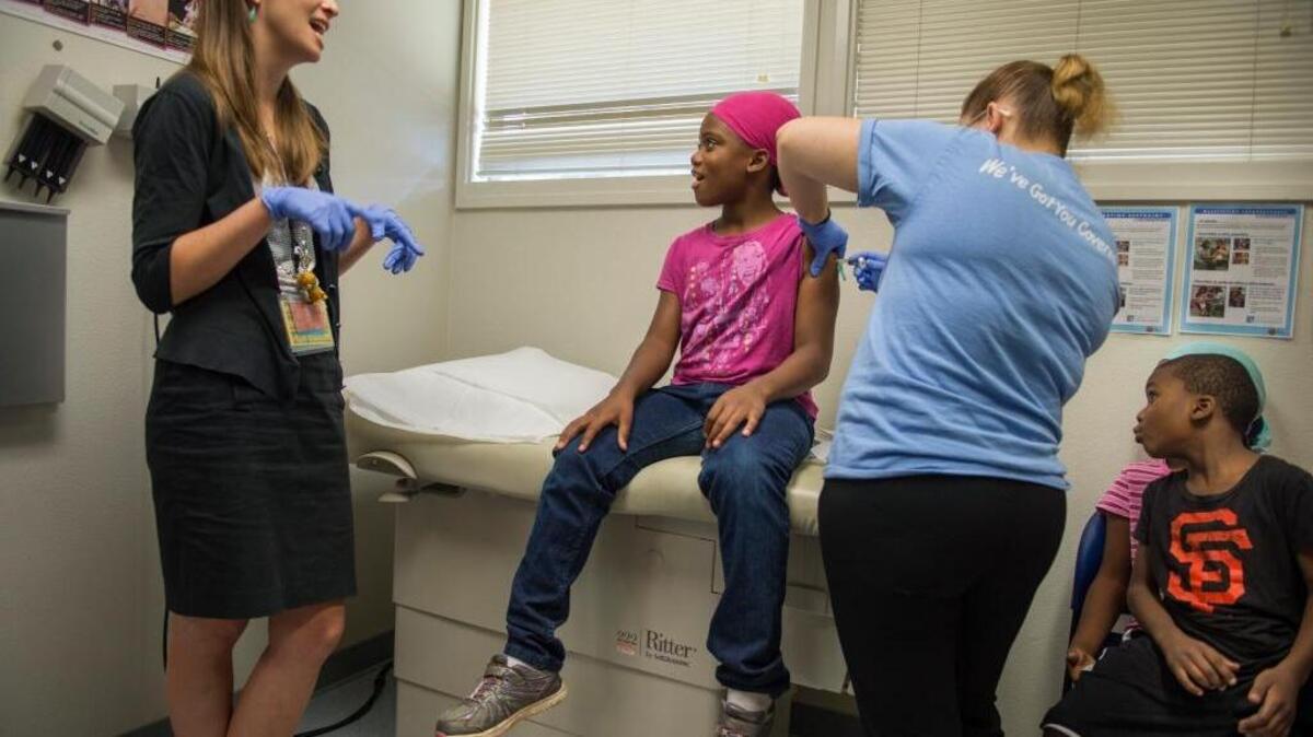 Emy Daniel, M.D., a resident pediatrician with UC Davis, left, distracts Lamaria Harris, 9, as her little brother, Neeko Harris, 5, right, gets a good look at her booster shot at the Oak Park Health Center in 2016. Since 2015, when California vaccine laws were tightened, students need current immunizations to begin the school year.
