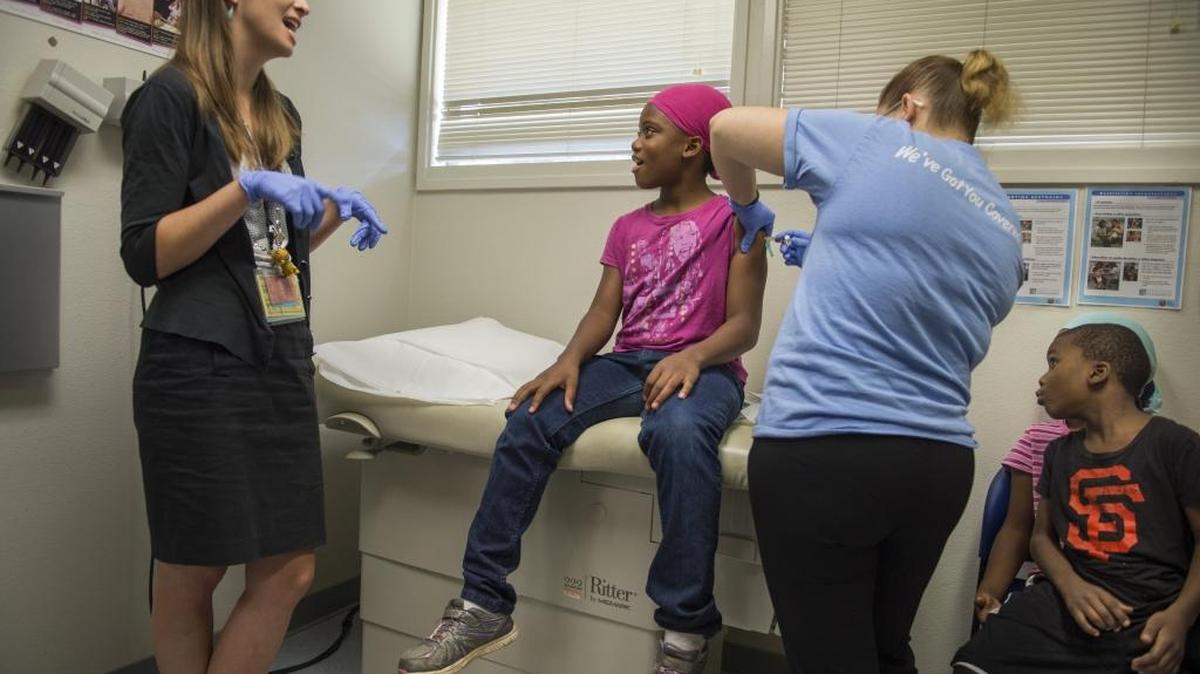 Dr. Emy Daniel, a pediatrician with UC Davis, provides distraction for Lamaria Harris, 9, as little brother Neeko Harris, 5, gets a good look at the booster shot administered by Marciela Magallon, a medical assistant with WellSpace Health in Oak Park. Due to new legislation, students who are not current on immunizations cannot begin the school year.